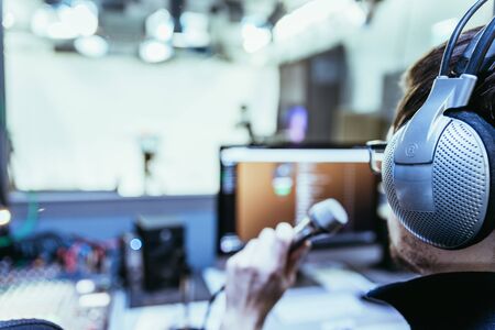 Young man is sitting in the recording studio and talks into the microphone, buttons and equipment in the blurry backgroundの写真素材