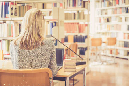 Blonde female student is sitting at the desk with pile of books, university library
