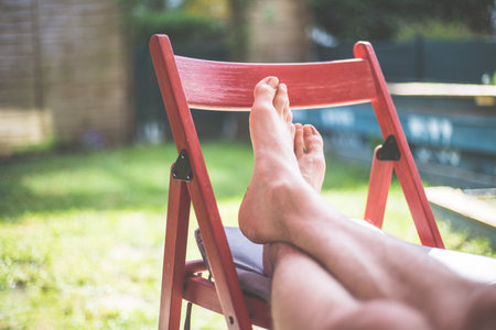 Young man is relaxing at home. Resting feet on a red chair, own garden in the blurry backgroundの写真素材