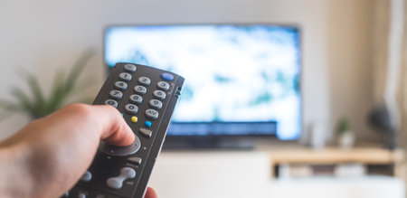Holding a TV remote control in the hand, foreground, tv in the blurry background. Streaming.の写真素材