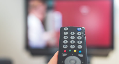 Holding a TV remote control in the hand, foreground, tv in the blurry background. Streaming.の写真素材