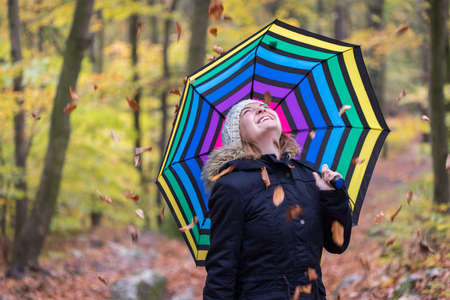Close up of pretty young girl with a colorful umbrella in autumnal forest. Leaves are falling down.の写真素材