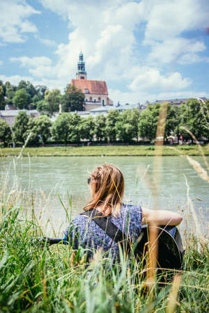 Beautiful young girl is sitting at a river and playing her western guitarの写真素材