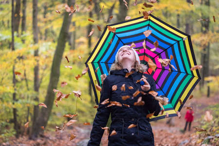 Close up of pretty young girl with a colorful umbrella in autumnal forest. Leaves are falling down.の写真素材