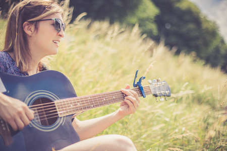 Beautiful young girl is sitting at a river and playing her western guitarの写真素材
