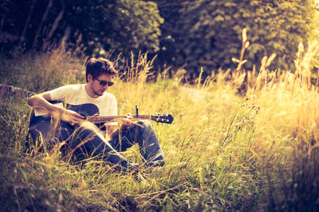 Young man is sitting at a river and playing his western guitarの写真素材