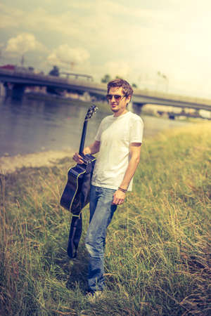 Young man is sitting at a river and playing his western guitarの写真素材