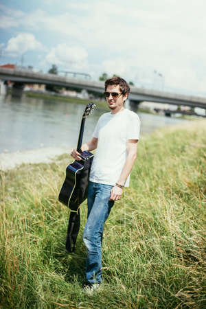 Young man is sitting at a river and playing his western guitarの写真素材