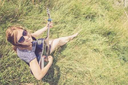 Beautiful young girl is sitting at a river and playing her western guitarの写真素材