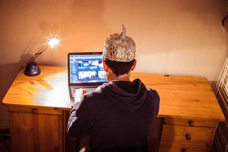 Young man with aluminum cap is sitting in the dark basement in front of a laptop. Conspiracy theory conceptの写真素材