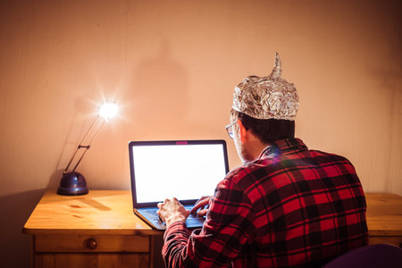 Young man with aluminum cap is sitting in the dark basement in front of a laptop. Conspiracy theory conceptの写真素材