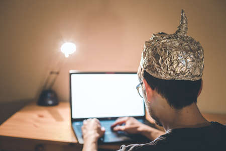 Young man with aluminum cap is sitting in the dark basement in front of a laptop. Conspiracy theory conceptの写真素材