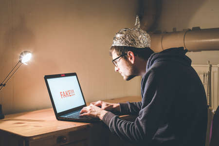 Young man with aluminum cap is sitting in the dark basement in front of a laptop. Conspiracy theory conceptの写真素材