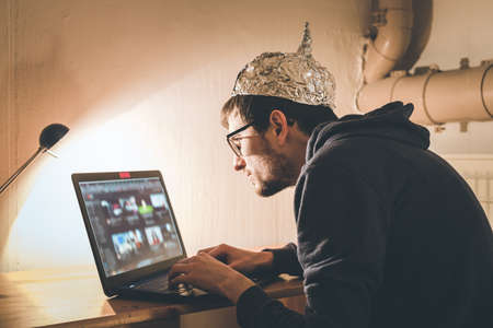 Young man with aluminum cap is sitting in the dark basement in front of a laptop. Conspiracy theory conceptの写真素材