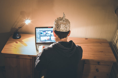 Young man with aluminum cap is sitting in the dark basement in front of a laptop. Conspiracy theory...