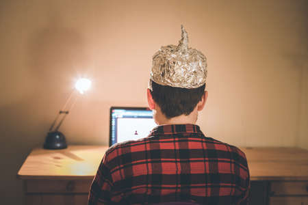 Young man with aluminum cap is sitting in the dark basement in front of a laptop. Conspiracy theory conceptの写真素材