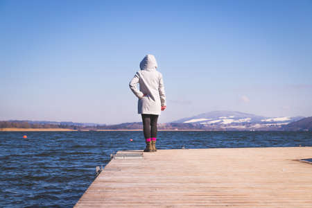 Young girl with gray coat is standing on a footbridge and enjoys the view over the lake, winter timeの写真素材
