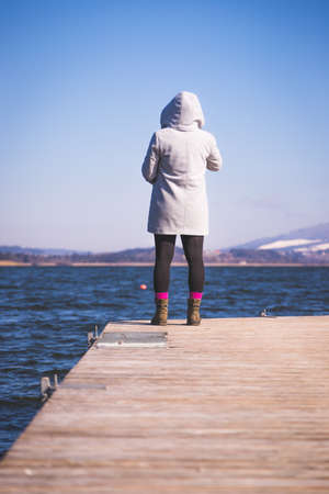 Young girl with gray coat is standing on a footbridge and enjoys the view over the lake, winter timeの写真素材