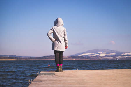 Young girl with gray coat is standing on a footbridge and enjoys the view over the lake, winter timeの写真素材