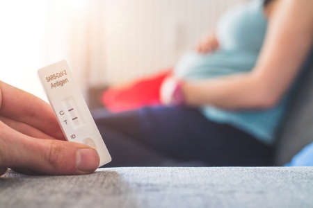 Close up of fingers holding express antigen covid test, positive result. Pregnant woman in the blurry backgroundの写真素材