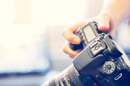 Photographer is holding a professional camera with telephoto lens in his hand, laptop in the blurry backgroundの写真素材