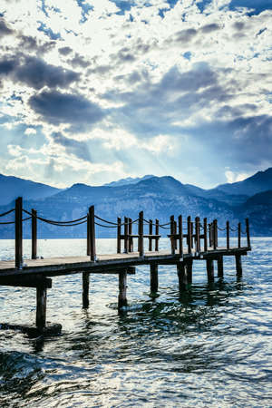 Wooden dock pier extending over blue lake water, mountains at lago di gardaの写真素材