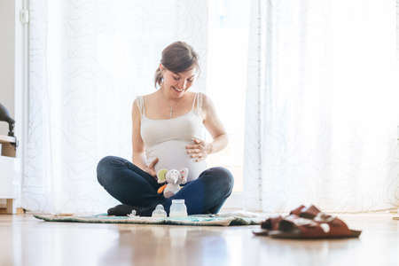 Happy Caucasian pregnant mother is sitting on the floor, touching her tummy, blue jeansの写真素材