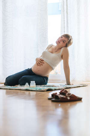 Happy Caucasian pregnant mother is sitting on the floor, touching her tummy, blue jeansの写真素材