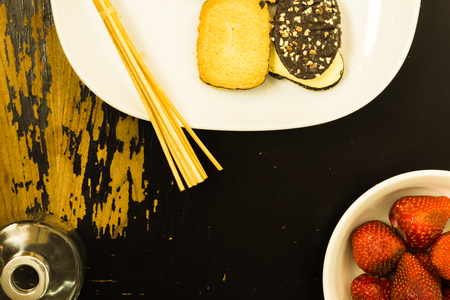 A plate of cookies, a glass jar and a bowl of strawberries are arranged on a scratchy wooden surfaceの写真素材
