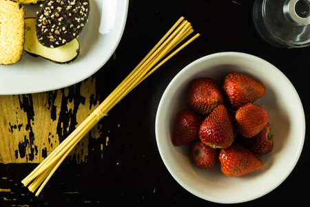 A plate of cookies, a glass jar and a bowl of strawberries are arranged on a scratchy wooden surfaceの写真素材