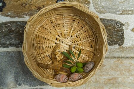 Fancy wicker basket on stone wall. Starfish, leaves and seashells inside.の写真素材