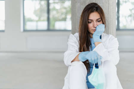 Sad and hopeless young female doctor holding protective mask for protecting from virus desease sitting on floor at hospital.の写真素材
