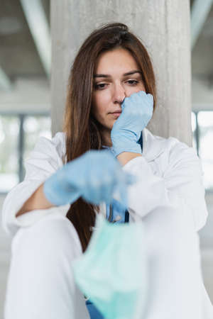 Sad and hopeless young female doctor holding protective mask for protecting from virus desease sitting on floor at hospital.の写真素材