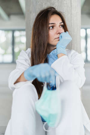 Sad and hopeless young female doctor holding protective mask for protecting from virus desease sitting on floor at hospital.の写真素材