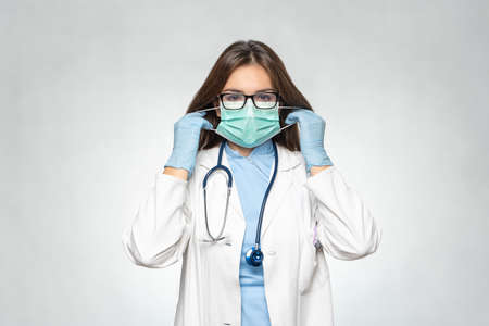 Confident young female doctor in white coat, latex protective gloves wearing medical mask. Woman in medical gloves put on protective mask. Coronavirus covid-19の写真素材