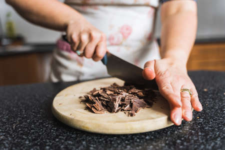 Woman chopping chocolate on wooden kitchen board in the kitchen wearing apron. Preparing ingredients for baking.の写真素材