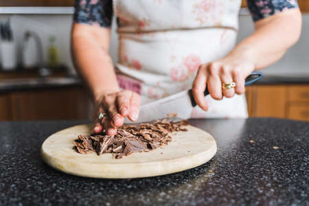 Woman chopping chocolate on wooden kitchen board in the kitchen wearing apron. Preparing ingredients for baking.の写真素材