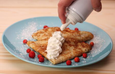detail of hand spraying whipped cream and two pancakes with wild strawberries  on  blue plate on wooden tableの写真素材