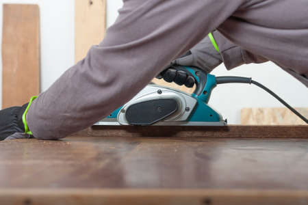 Carpenter working with electric planer on wood lath on desk, on background oriented strand board