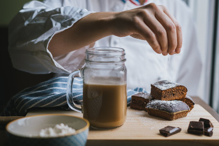 Chef with a white apron decorating brownies with flour on a wooden board with a cup of coffee with milk, ounces of chocolate and a blue cloth.の写真素材