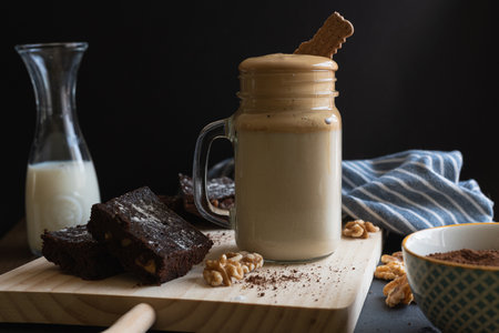Dalgona coffee cup with a cookie on top on a wooden board with brownies and nuts, a jug of milk and a black background.の写真素材