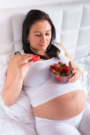 Beautiful pregnant woman with a bowl of strawberries in bedの写真素材