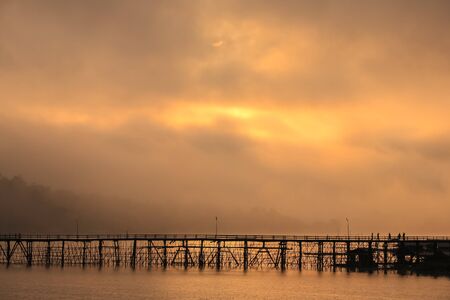 Wooden bridge across through river with foggy background at sunrise reflection on water located Kanchanaburee province west of Thailandの写真素材