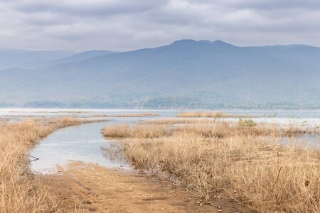Dry land of field under cloudy sky and mountain in front of water in damの写真素材