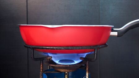 close-up of hand holding red frying pan on retro and old rustic gas stove with flame on stove top for cooking.の写真素材