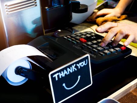cashier and cash register at counter with hand to poke numeric keypad to bill payment and thank you on blackboard sign.の写真素材