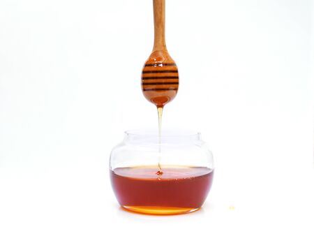 Drop of honey from wooden honey stick into clear glass jar isolated on white background.の写真素材