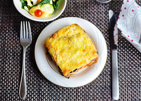 A piece of eggplant lasagna on a white dish with knife, fork, and salad (above)の写真素材