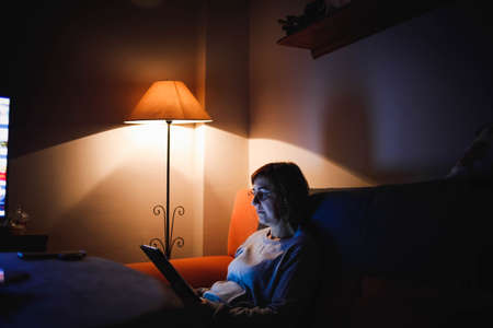 adult woman using a touch tablet in the living room. Adult woman using a touch device. Woman reading a book. Woman making video call with familyの写真素材