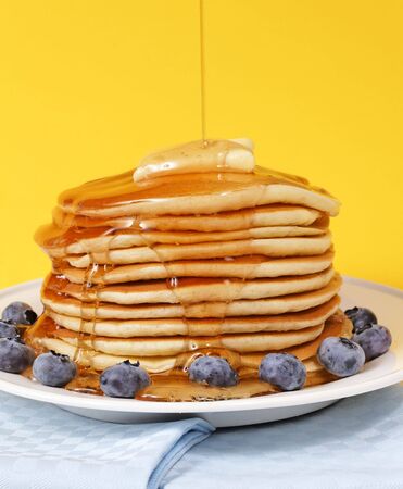 Hot pancake staked with blueberry fruit on a yellow background. Melted butter on the top with maple syrup falling on it.の写真素材